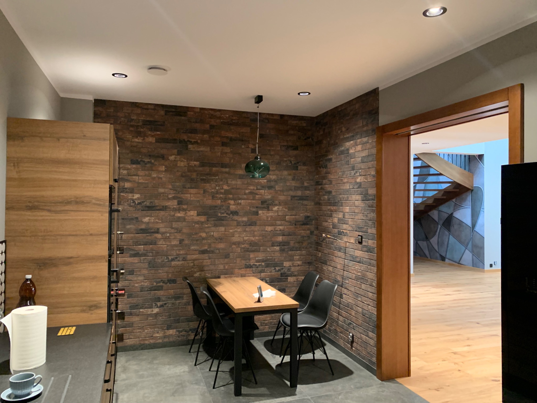 Jadalnia z ceglaną ścianą, drewnianym stołem i czarnymi krzesłami. W tle widoczne schody i parkiet.