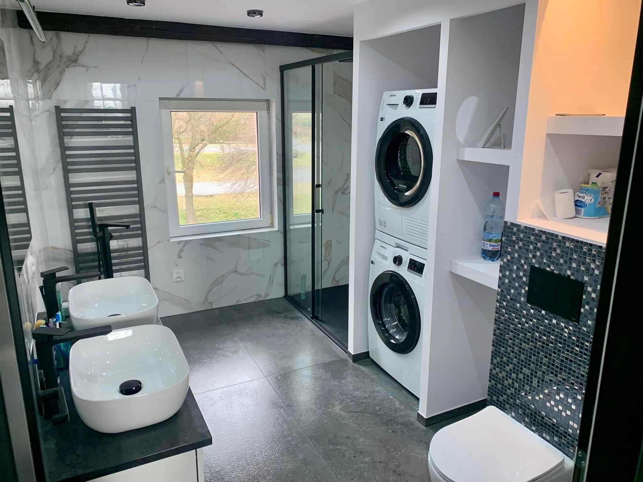 Nowoczesna łazienka z dwoma umywalkami nablatowymi, kabiną prysznicową z czarnymi ramami, pralką i suszarką w zabudowie oraz toaletą z mozaiką ścienną.