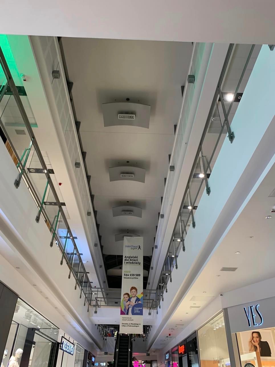 Wnętrze galerii handlowej widziane z dołu, z widocznymi balustradami, oświetleniem sufitowym oraz banerem reklamowym szkoły językowej Helen Doron English, wiszącym nad schodami ruchomymi.
