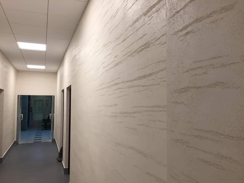 Korytarz z dekoracyjną, jasną ścianą o fakturze przypominającej piaskowiec z delikatnymi, poziomymi smugami, widoczne drzwi w głębi.