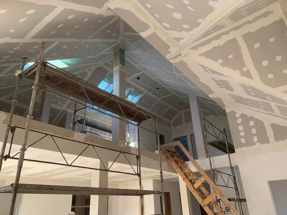 Wysokie wnętrze w trakcie prac wykończeniowych: ściany i sufit pokryte płytami gipsowo-kartonowymi, widoczne szpachlowanie łączeń, rusztowania i drabina. W tle okna dachowe wpadające do wnętrza.