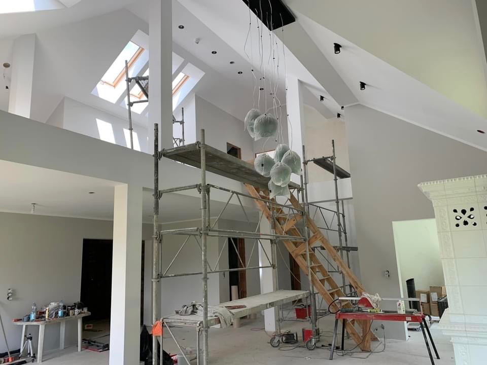 Wnętrze domu w trakcie malowania: widoczne rusztowania, zabezpieczone folią lampy, pomalowane ściany na biało, okna dachowe i narzędzia malarskie.