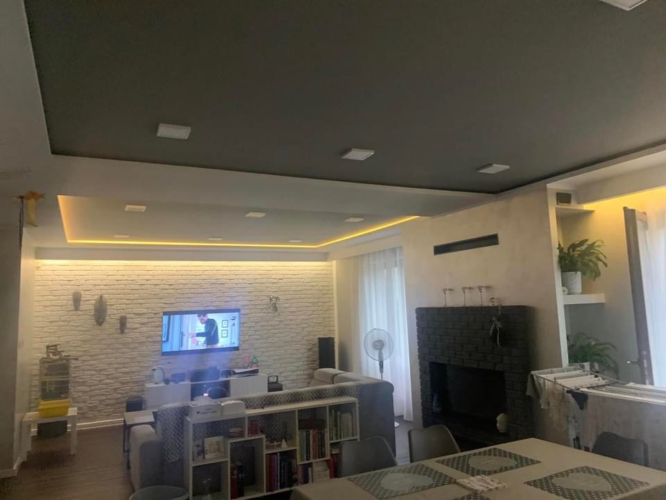 Salon z ceglaną ścianą, podwieszanym sufitem z oświetleniem LED, kominkiem z ciemnej cegły i roślinami na półkach.