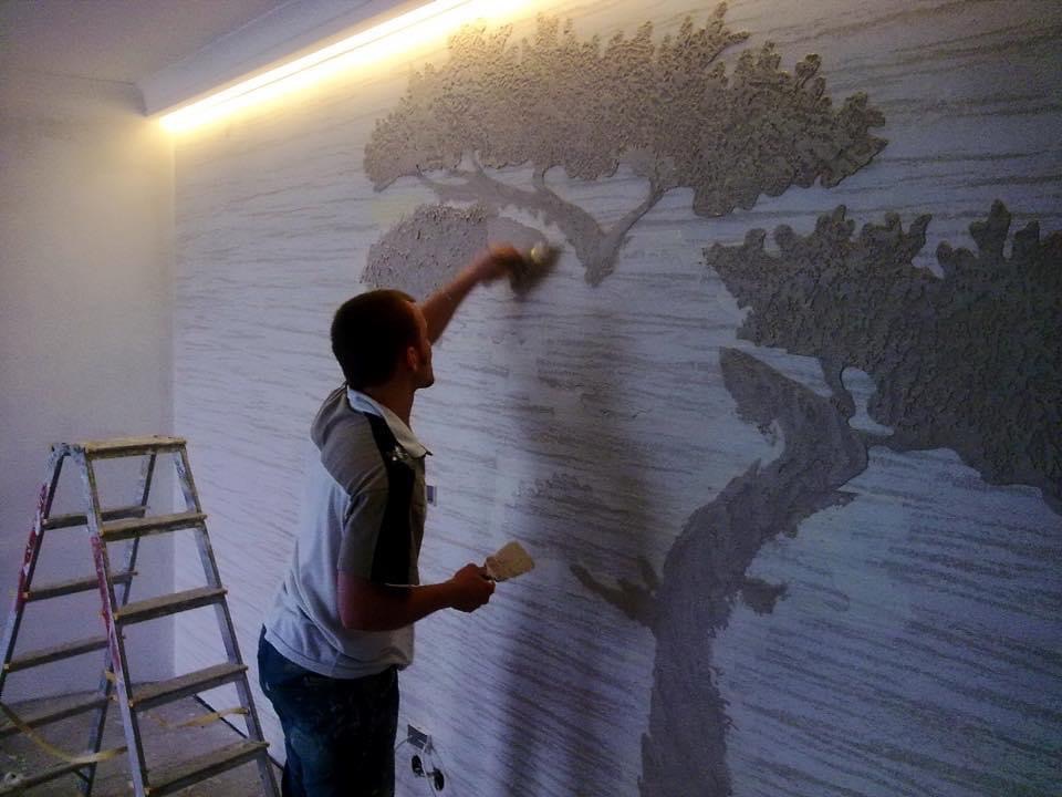 Mężczyzna aplikuje dekoracyjny tynk strukturalny w kształcie drzewa na ścianie w pokoju, drabina stoi obok.