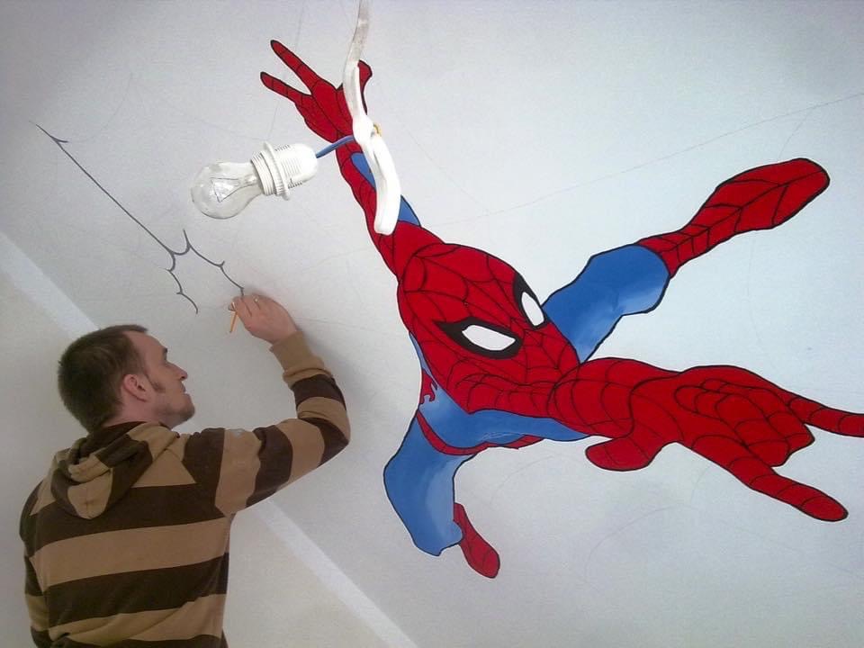 Mężczyzna w bluzie w paski rysuje ołówkiem pajęczynę na suficie obok niedokończonego malunku Spider-Mana, z żarówką zwisającą z sufitu.