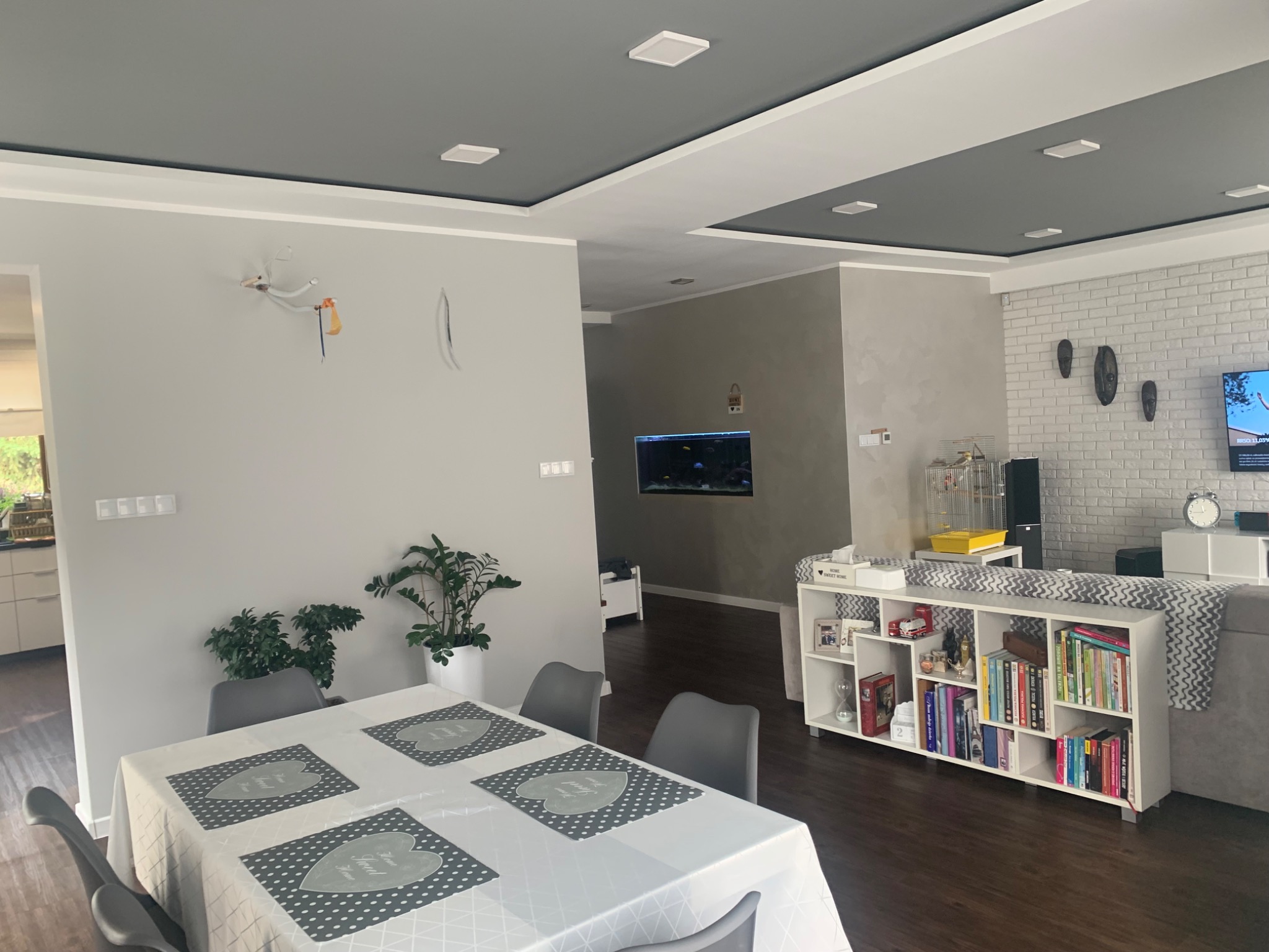 Jasne wnętrze salonu z pomalowanym na szaro sufitem, białymi ścianami, akwarium w ścianie i widocznym okablowaniem po demontażu lampy na ścianie.