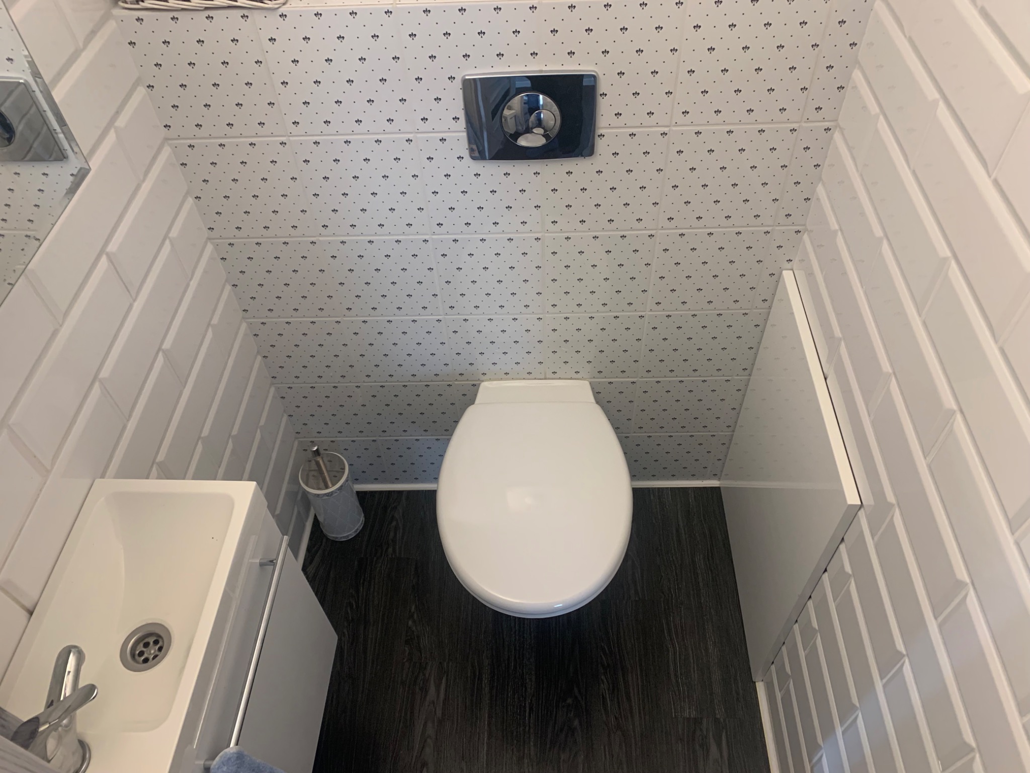 Nowoczesna toaleta z podwieszaną miską WC, umywalką narożną i ciemną podłogą imitującą drewno; ściany wyłożone białymi, prostokątnymi płytkami oraz płytkami z drobnym wzorem.