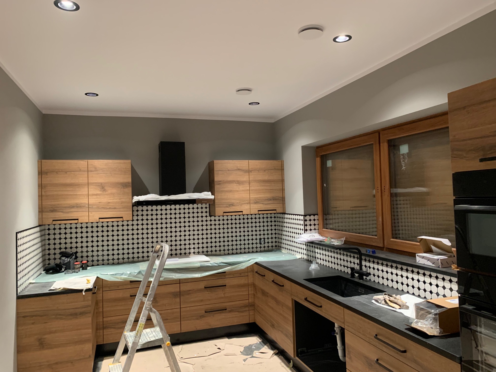 Kuchnia w trakcie wykańczania: drewniane szafki, czarny blat, mozaika na ścianie, widoczna drabina malarska i zabezpieczenia na blacie.