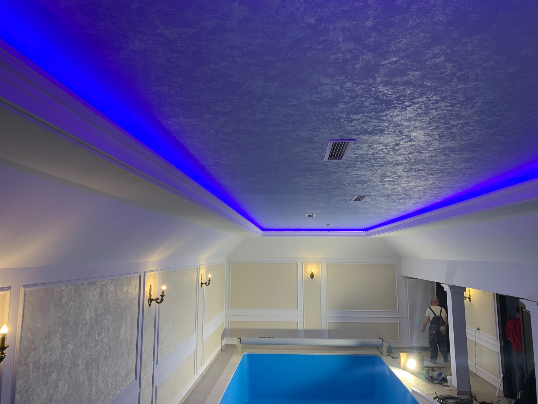 Wnętrze basenu z dekoracyjnym oświetleniem LED podsufitowym i ścianami wykończonymi w stylu klasycznym, widoczny pracownik w kombinezonie na tle kolumn.