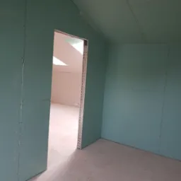 Wnętrze poddasza z zamontowanymi zielonymi płytami gipsowo-kartonowymi na ścianach i suficie, widoczne wkręty mocujące, surowy stan przed wykończeniem, widok na sąsiednie pomieszczenie przez otw&oacute;r...