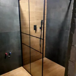 Nowoczesna kabina prysznicowa walk-in z czarnymi profilami, zamontowana w łazience z szarymi płytkami na ścianach i drewnopodobnymi płytkami na podłodze, widoczne przyłącza wodne.