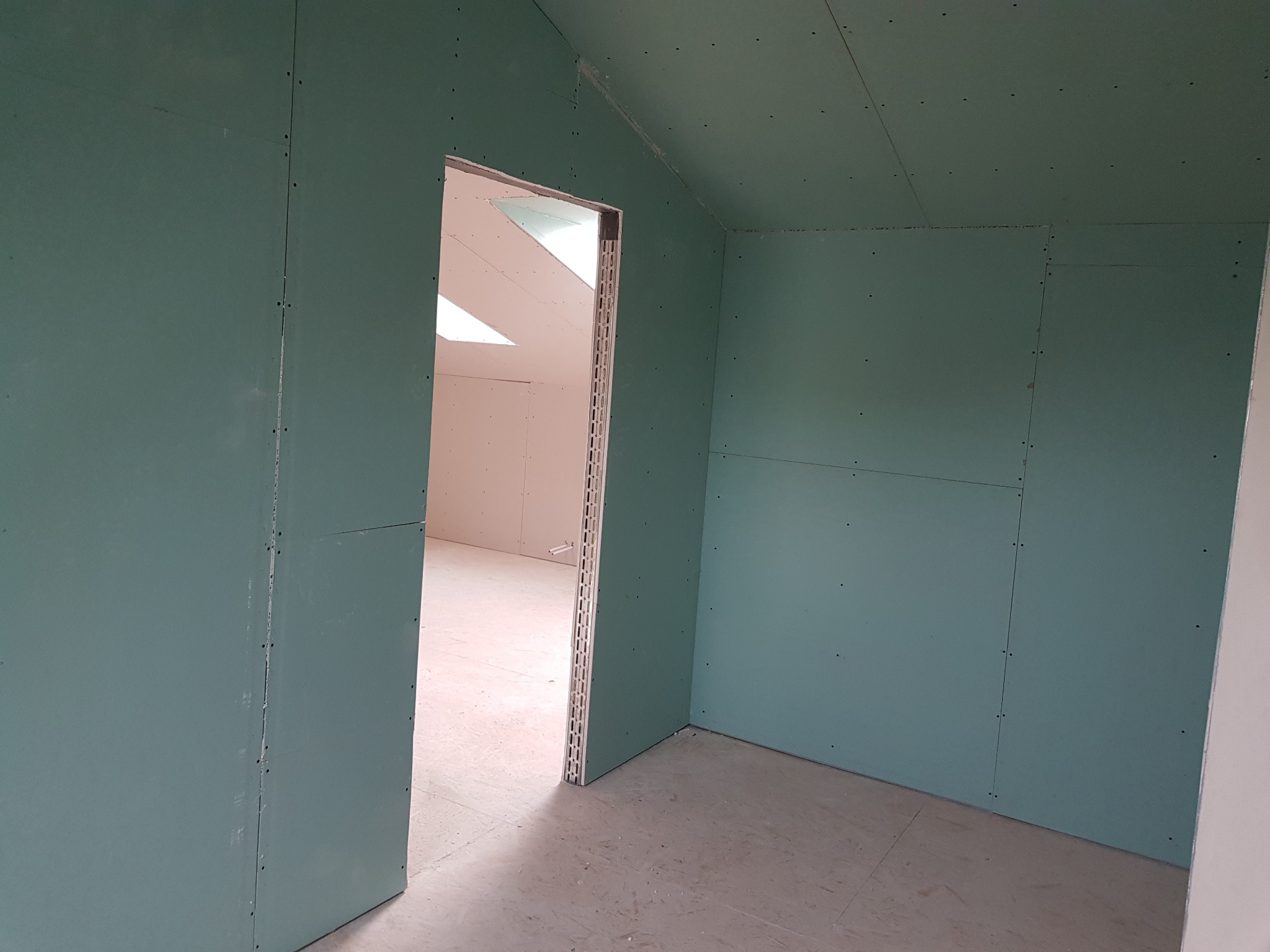 Wnętrze poddasza z zamontowanymi zielonymi płytami gipsowo-kartonowymi na ścianach i suficie, widoczne wkręty mocujące, surowy stan przed wykończeniem, widok na sąsiednie pomieszczenie przez otwór...