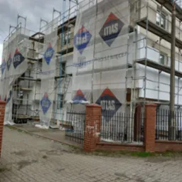 Wielopiętrowy budynek w trakcie prac, osłonięty siatką z logo Atlas, widoczne rusztowania i fragmenty ocieplenia na ścianach, ogrodzenie z ceglanymi słupkami w tle.