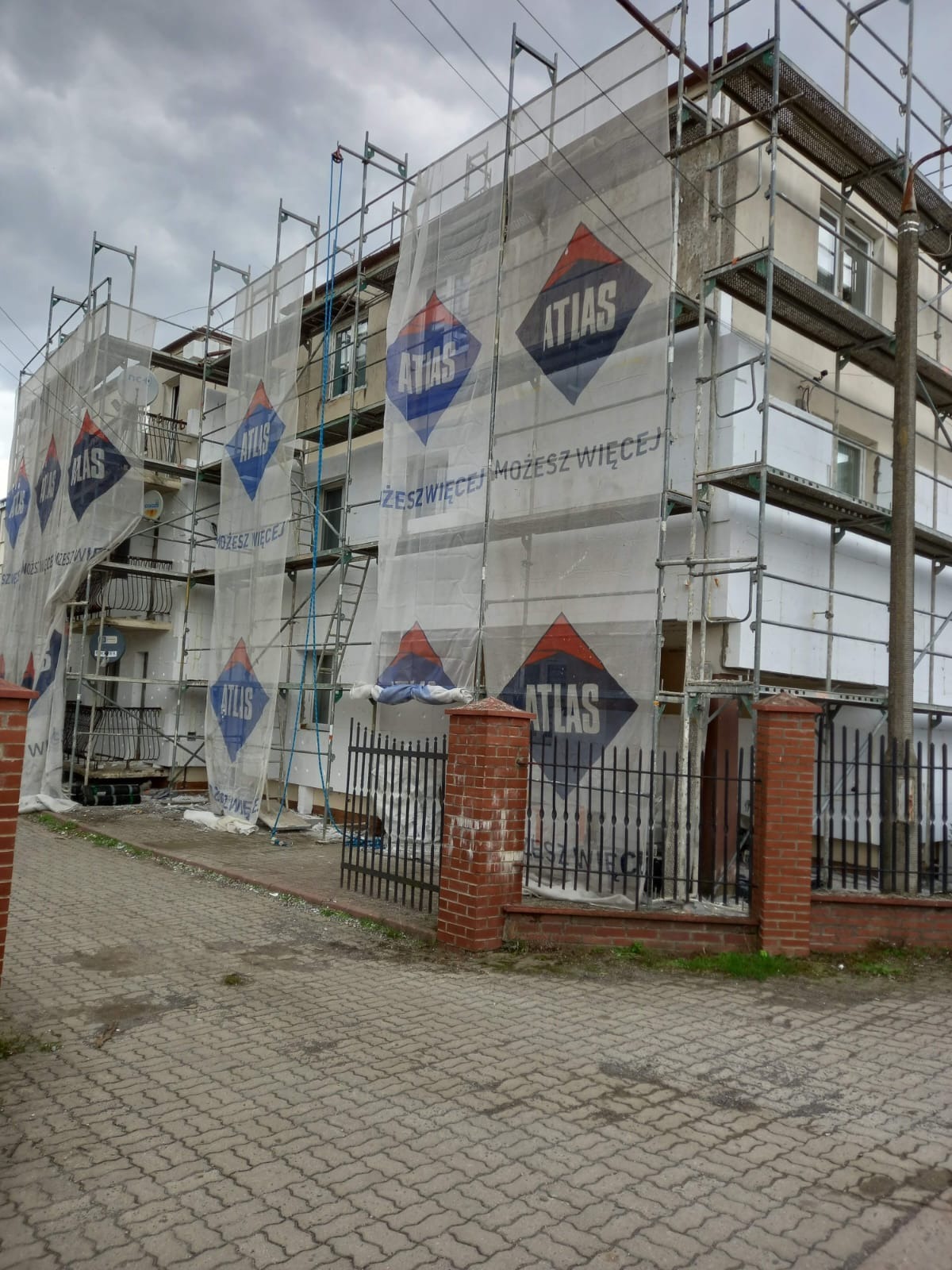Wielopiętrowy budynek w trakcie prac, osłonięty siatką z logo Atlas, widoczne rusztowania i fragmenty ocieplenia na ścianach, ogrodzenie z ceglanymi słupkami w tle.