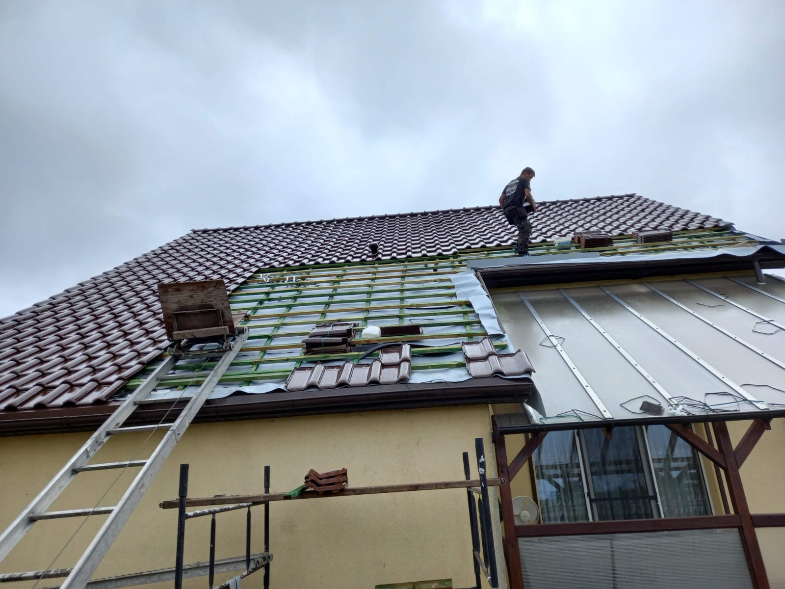 Prace dekarskie w toku: robotnik na dachu krytym częściowo brązową dachówką, widoczne drewniane łaty, drabina z podnośnikiem do dachówek, fragment dobudówki z poliwęglanu, pochmurne niebo.