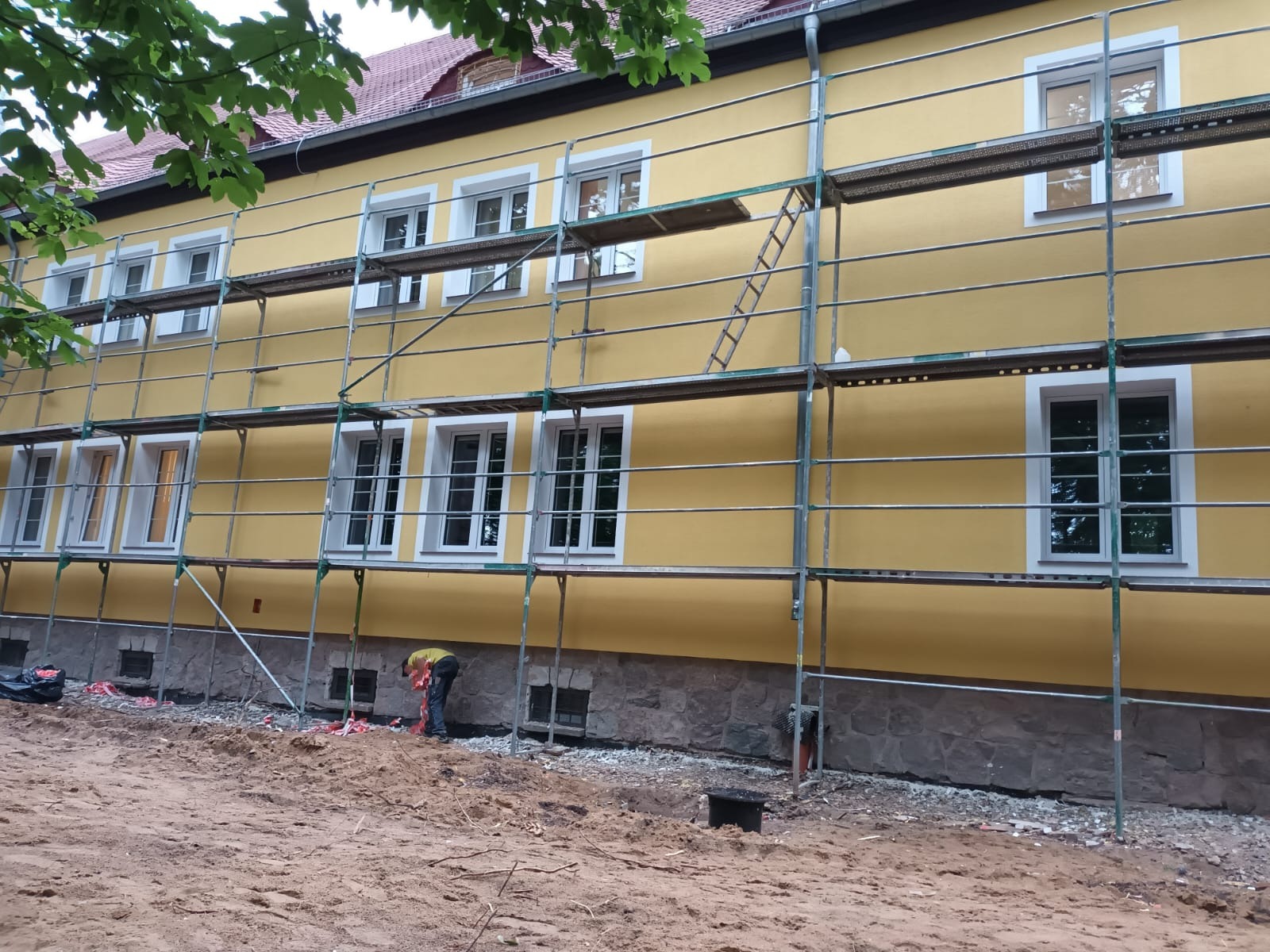 Żółta elewacja budynku w trakcie prac, widoczne rusztowania, pracownik przy ziemi, podmurówka z kamienia. Ujęcie z dołu, widoczne fragmenty drzewa w górnym rogu.