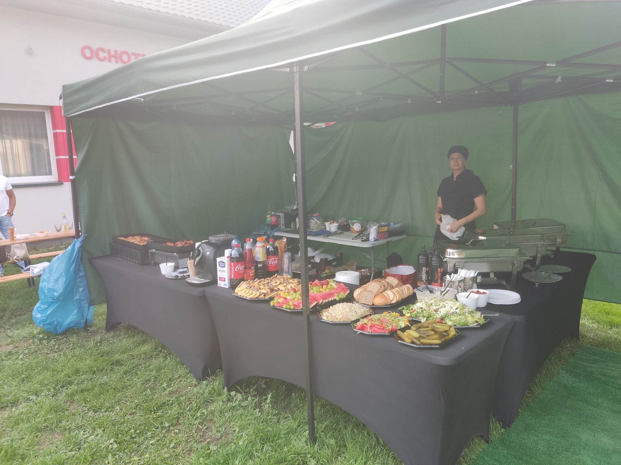 Stoisko cateringowe pod zielonym namiotem z różnorodnym jedzeniem: grillowane mięso, owoce, pieczywo, sałatki i napoje, obsługiwane przez osobę w czarnym uniformie.