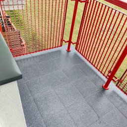 Sławomir Wołkowicz Flex Remonty - Szare płytki na balkonie z czerwoną, metalową balustradą. Widok z góry na wykończony balkon z widokiem na zieleń. Nowoczesne wykończenie balkonu.