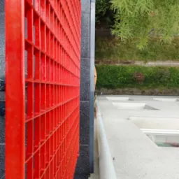 Czerwona, metalowa balustrada balkonu z widokiem na zieleń i elewację budynku. Nietypowy kadr z g&oacute;ry, podkreślający geometrię i kontrast kolor&oacute;w.