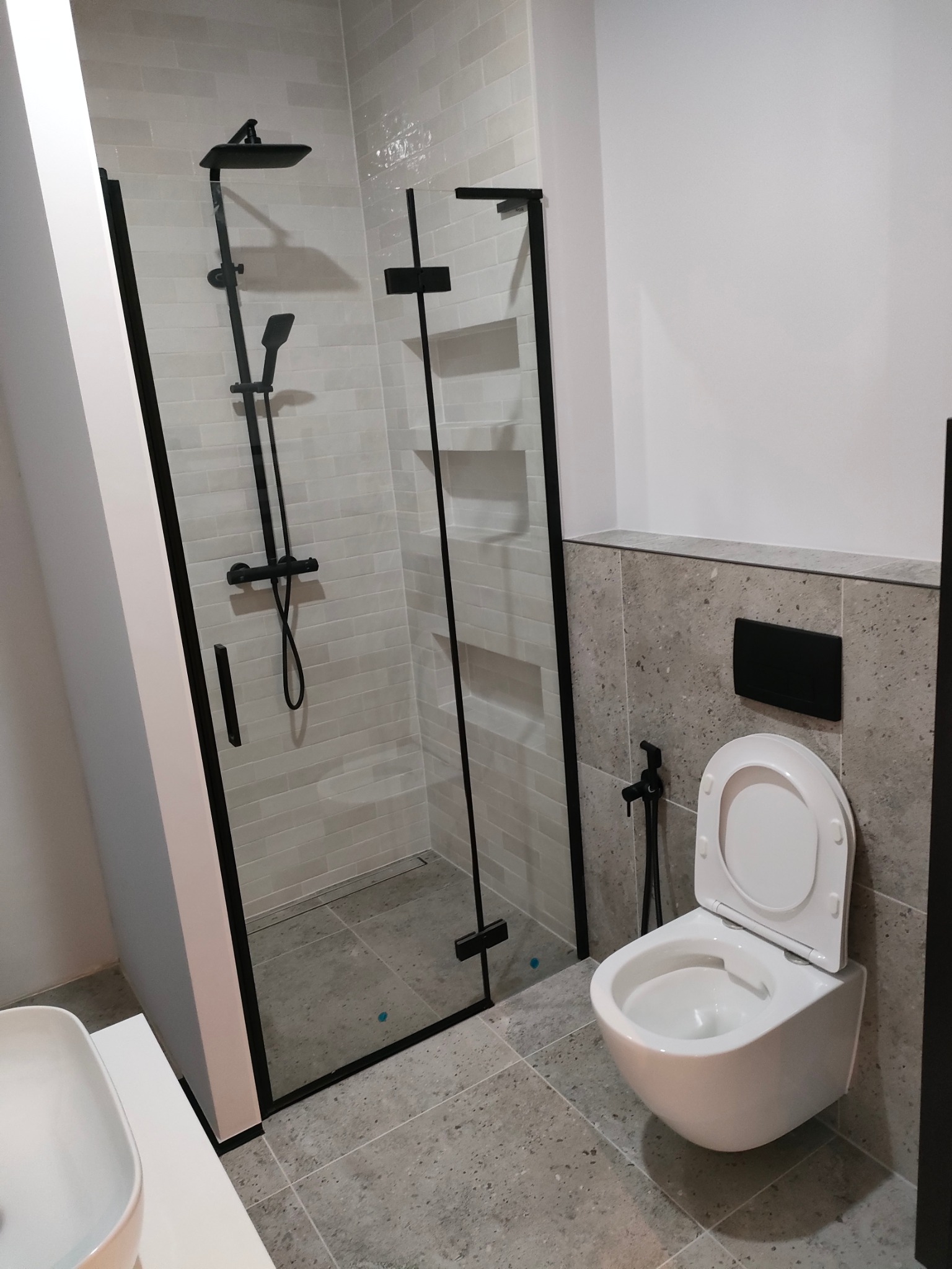 Nowoczesna łazienka z kabiną prysznicową typu walk-in z czarnymi profilami i armaturą, podwieszana toaleta z bidetką, szare płytki imitujące kamień.
