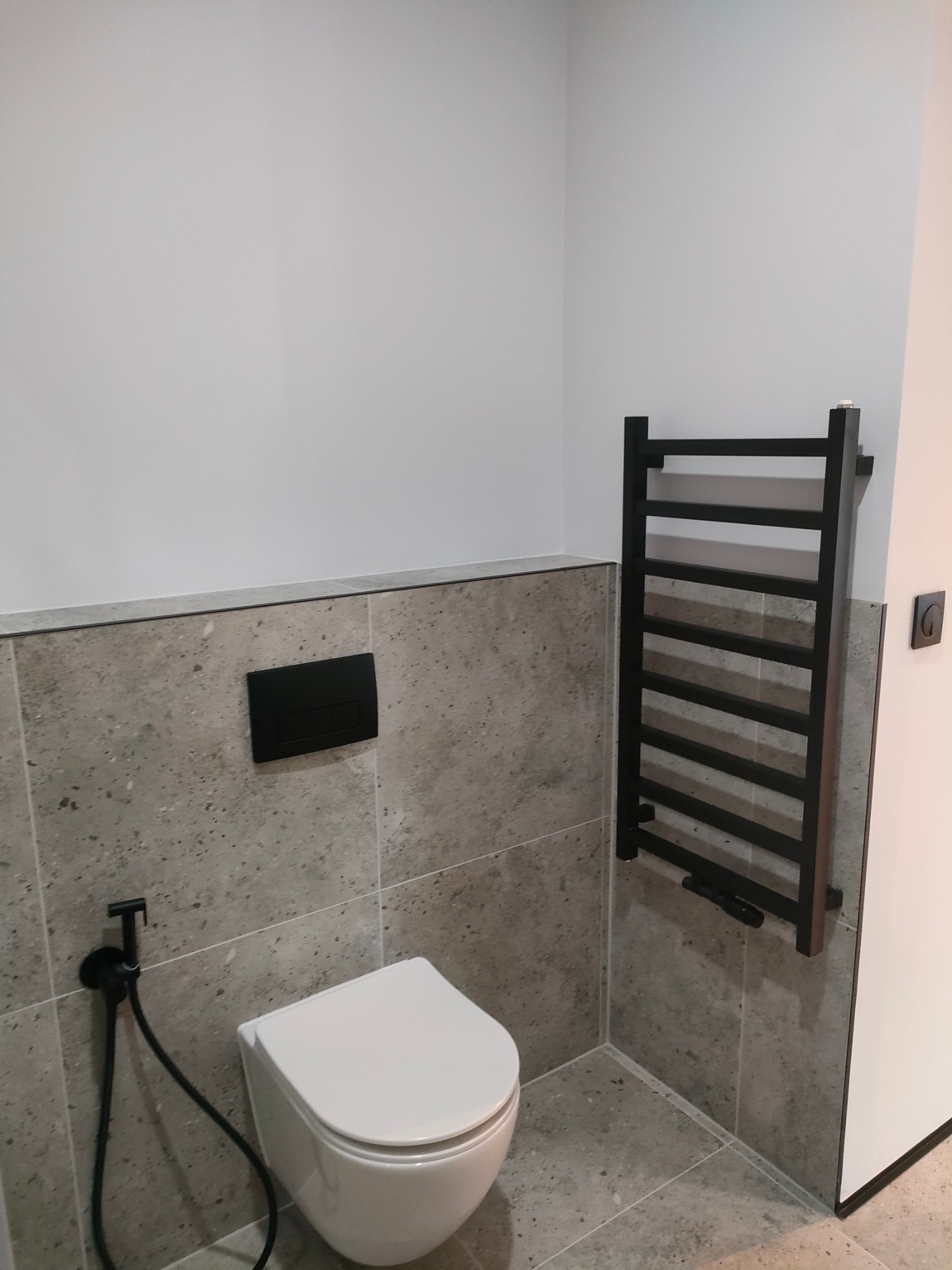 Nowoczesna łazienka z szarą, kamienną okładziną ścian, wiszącą toaletą z czarnym panelem sterującym, czarnym bidetem i grzejnikiem drabinkowym w kolorze czarnym.