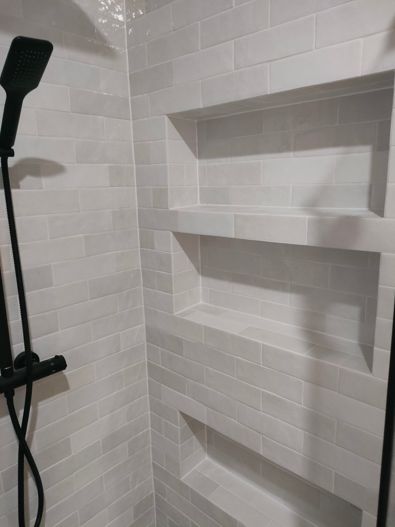 Nowoczesna kabina prysznicowa z białymi, podłużnymi płytkami i wnękami w ścianie, czarna bateria prysznicowa.