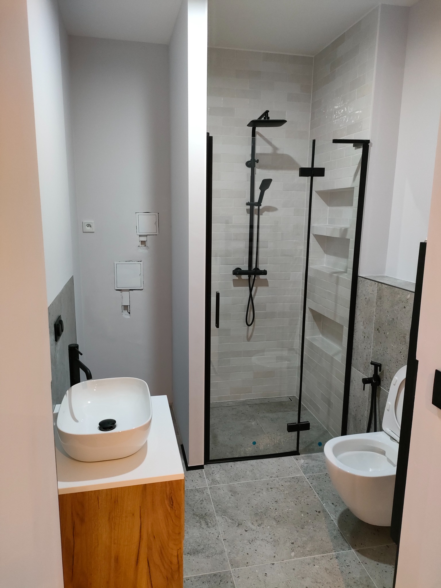 Nowoczesna łazienka z kabiną prysznicową typu walk-in z czarnymi elementami, umywalką nablatową na drewnianej szafce i wiszącą toaletą, wykończona w szarościach.