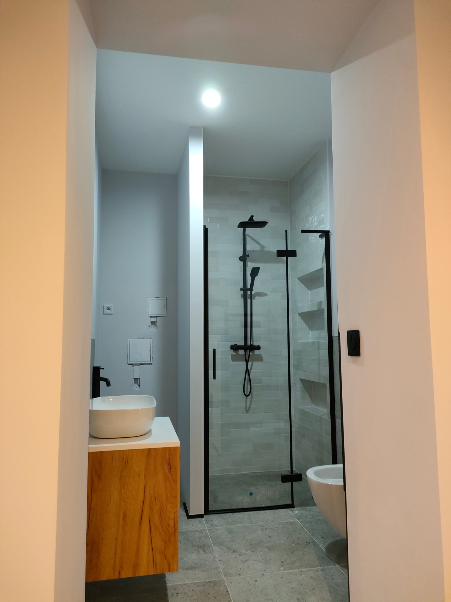 Nowoczesna łazienka z kabiną prysznicową typu walk-in z czarnymi profilami, umywalką nablatową na drewnianej szafce i szarą płytką podłogową.