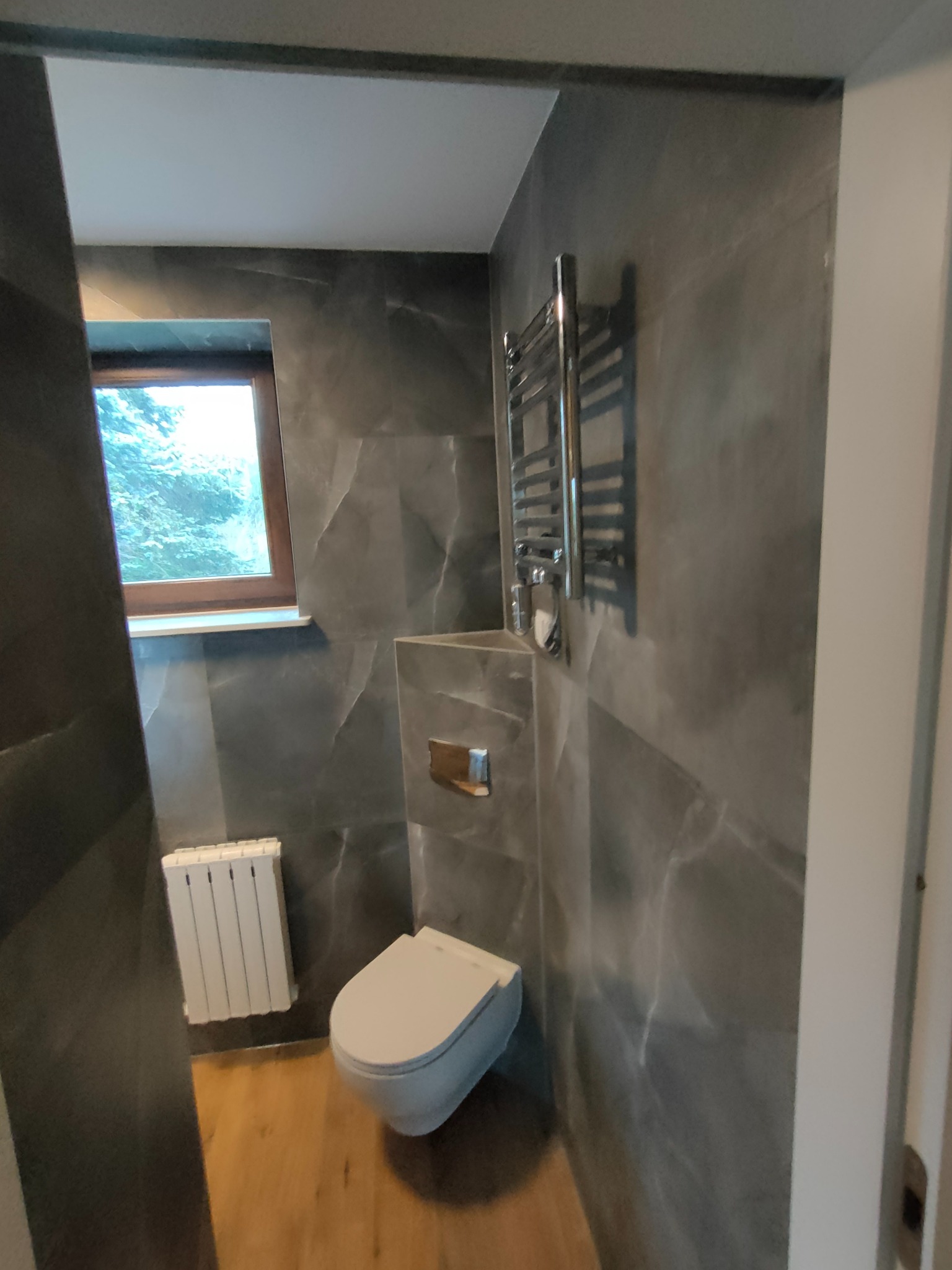 Nowoczesna łazienka z szarymi płytkami imitującymi marmur, wisząca toaleta, grzejnik drabinkowy i drewniana podłoga, widok z perspektywy wejścia.