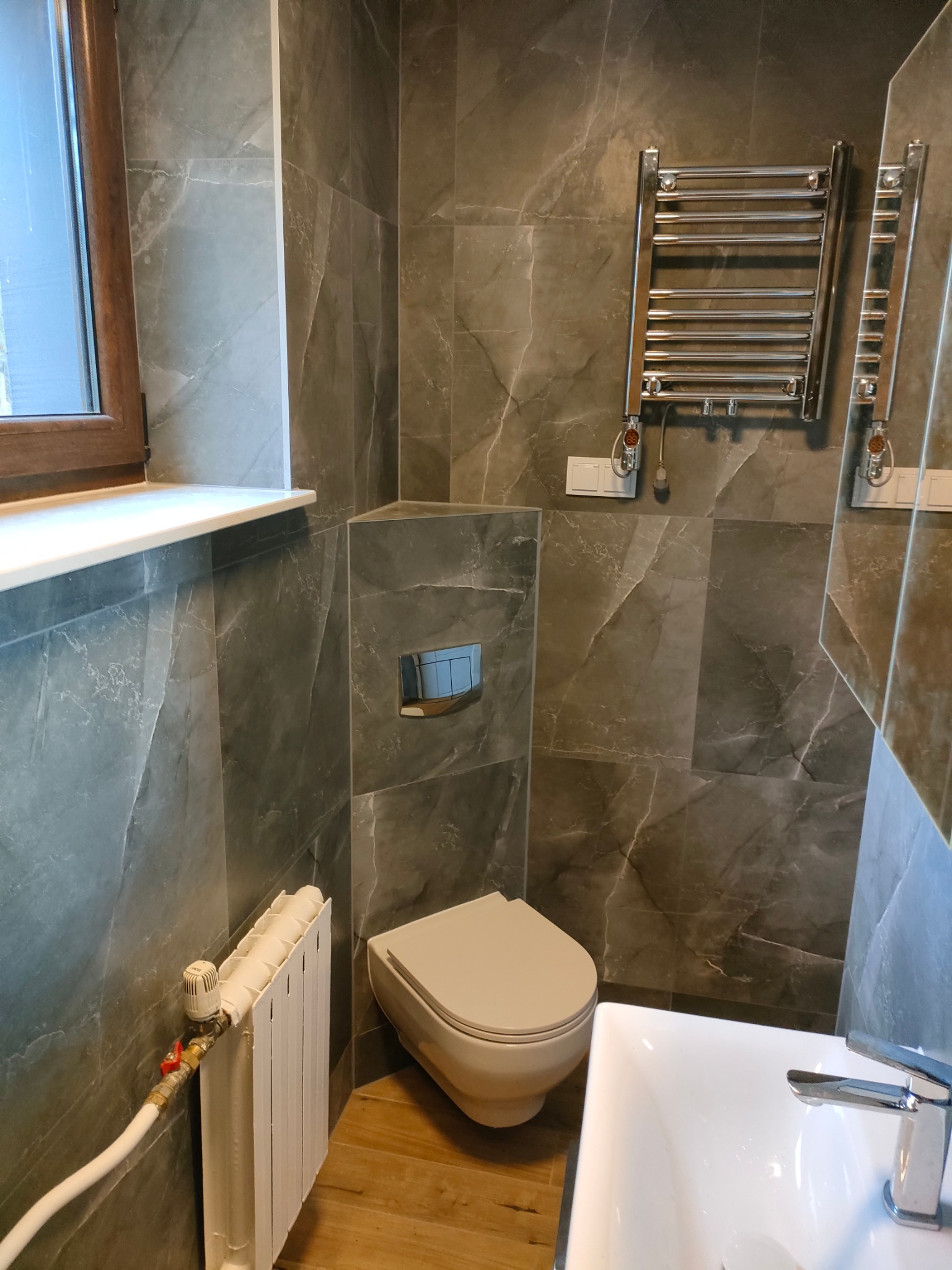 Wykończona łazienka z szarymi płytkami imitującymi marmur, wiszącą muszlą klozetową, grzejnikiem drabinkowym i umywalką nablatową.