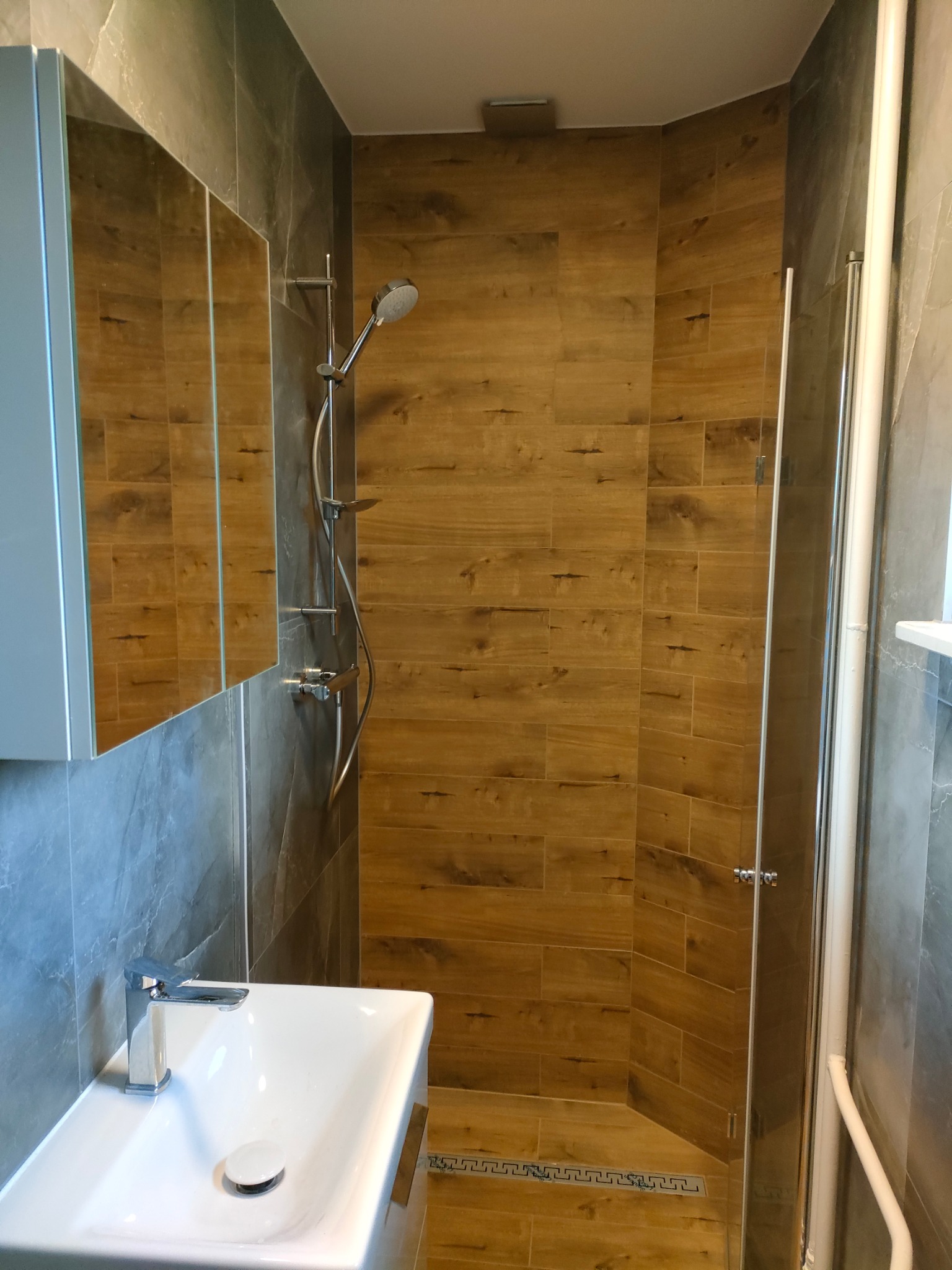 Nowoczesna kabina prysznicowa z deszczownicą i odpływem liniowym wykończona płytkami imitującymi drewno, umywalka nablatowa z chromowaną baterią, szare płytki na ścianach