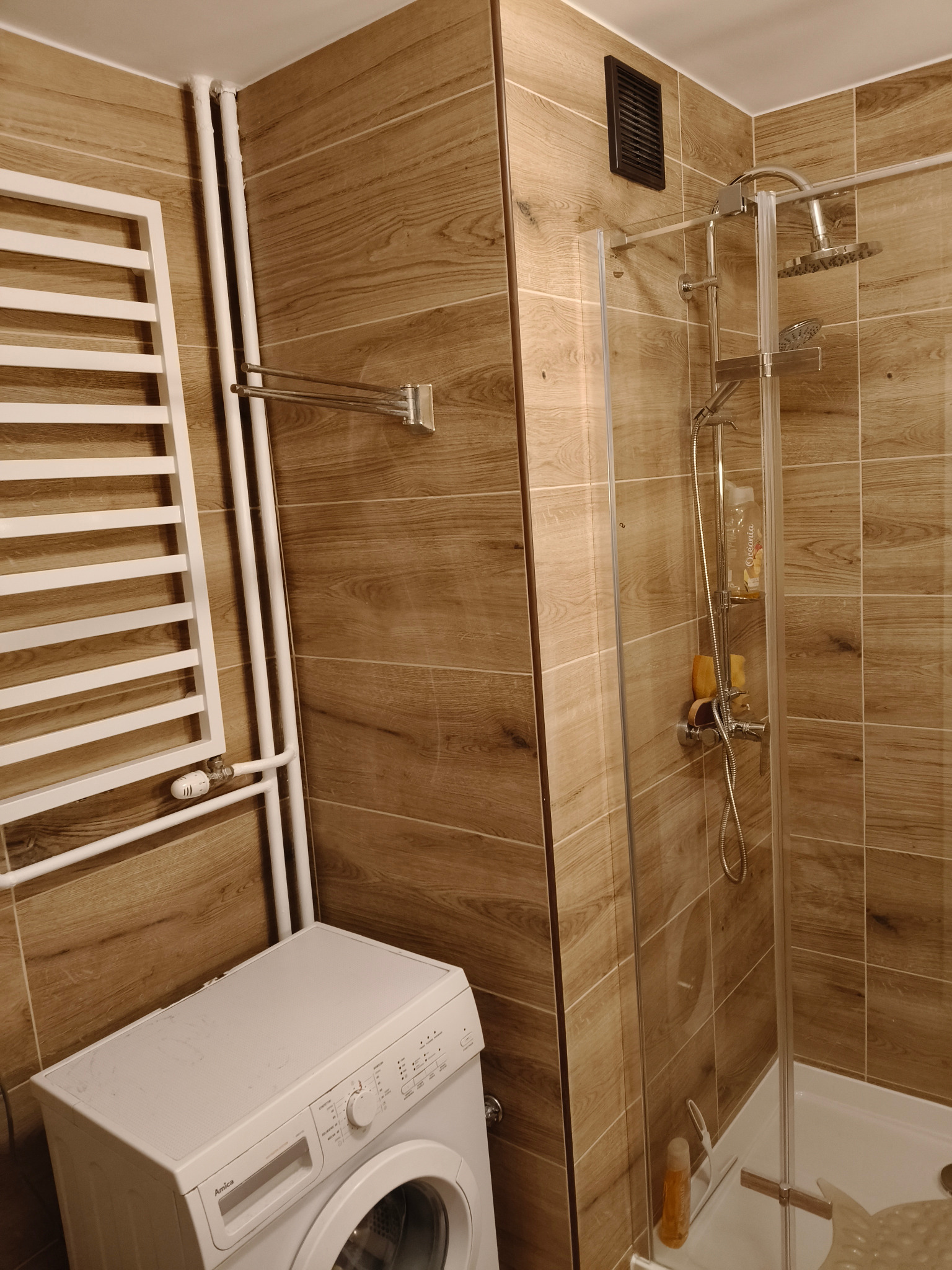 Ukończona łazienka z kabiną prysznicową, grzejnikiem drabinkowym i pralką. Płytki imitujące drewno na ścianach. Nowoczesny design.