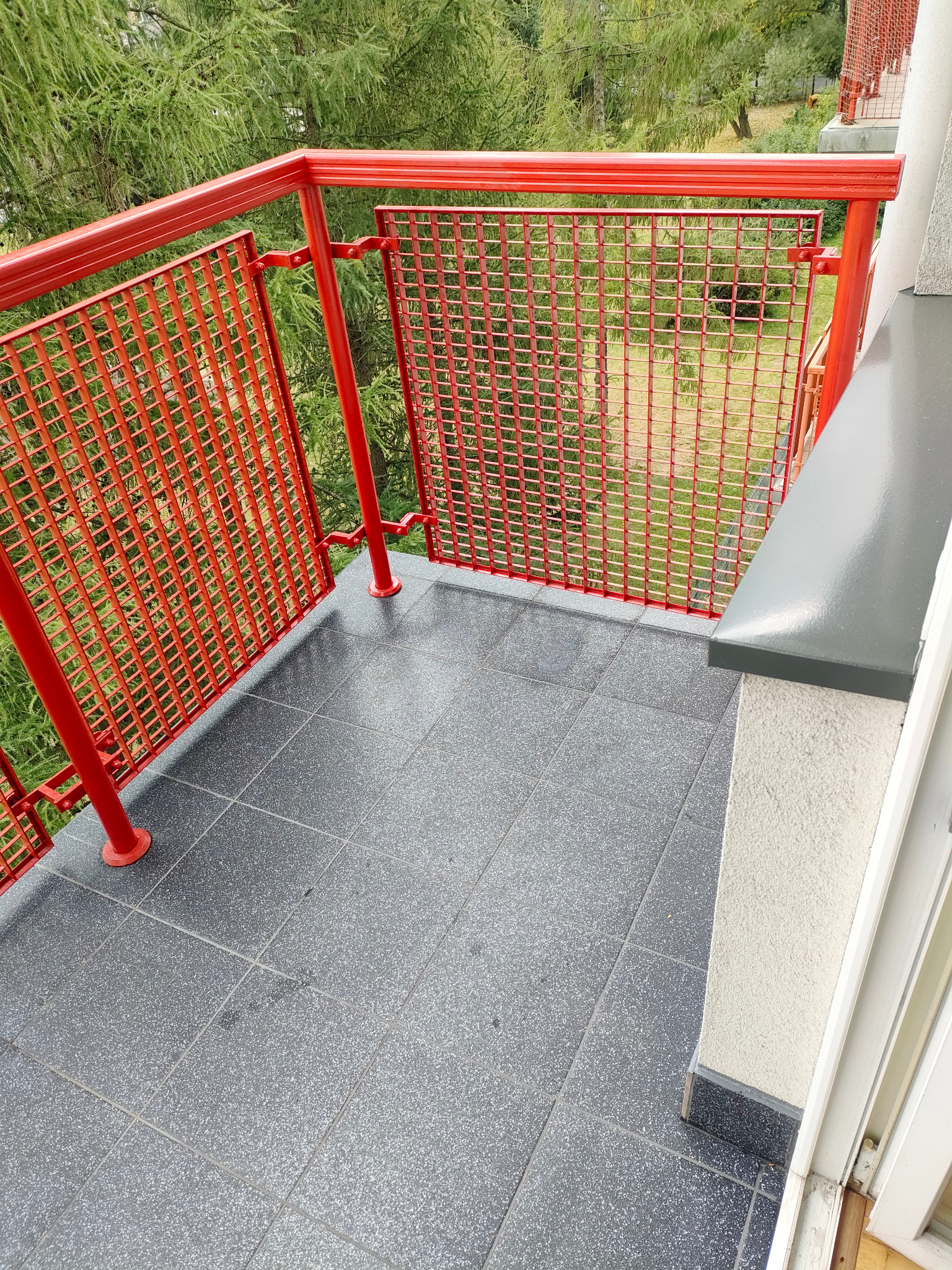 Wykończony balkon z szarą, nakrapianą podłogą z płytek i czerwoną, metalową balustradą. Widok na zieleń zza balustrady. Świeżo położone płytki.