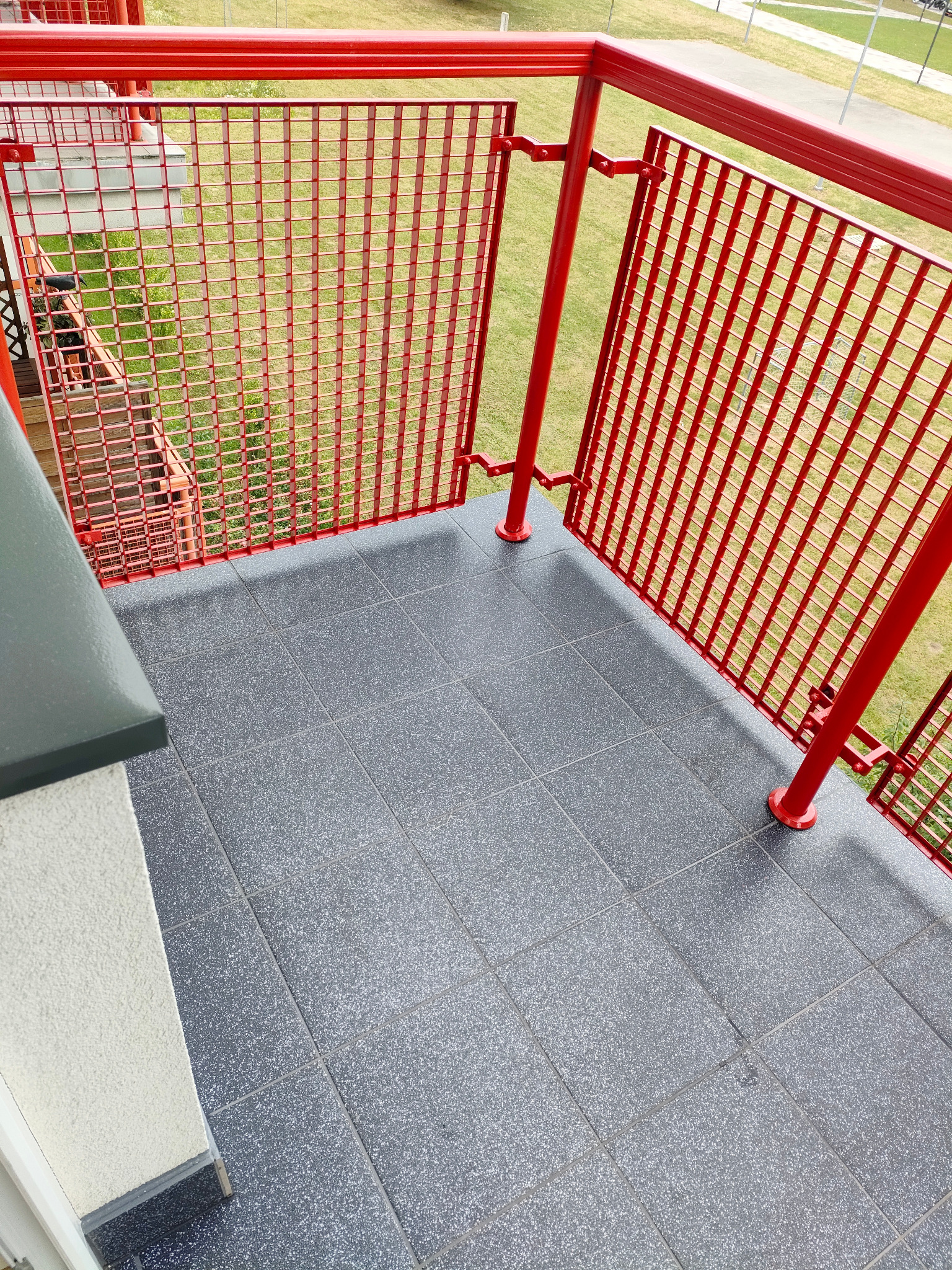 Szare płytki na balkonie z czerwoną, metalową balustradą. Widok z góry na wykończony balkon z widokiem na zieleń. Nowoczesne wykończenie balkonu.