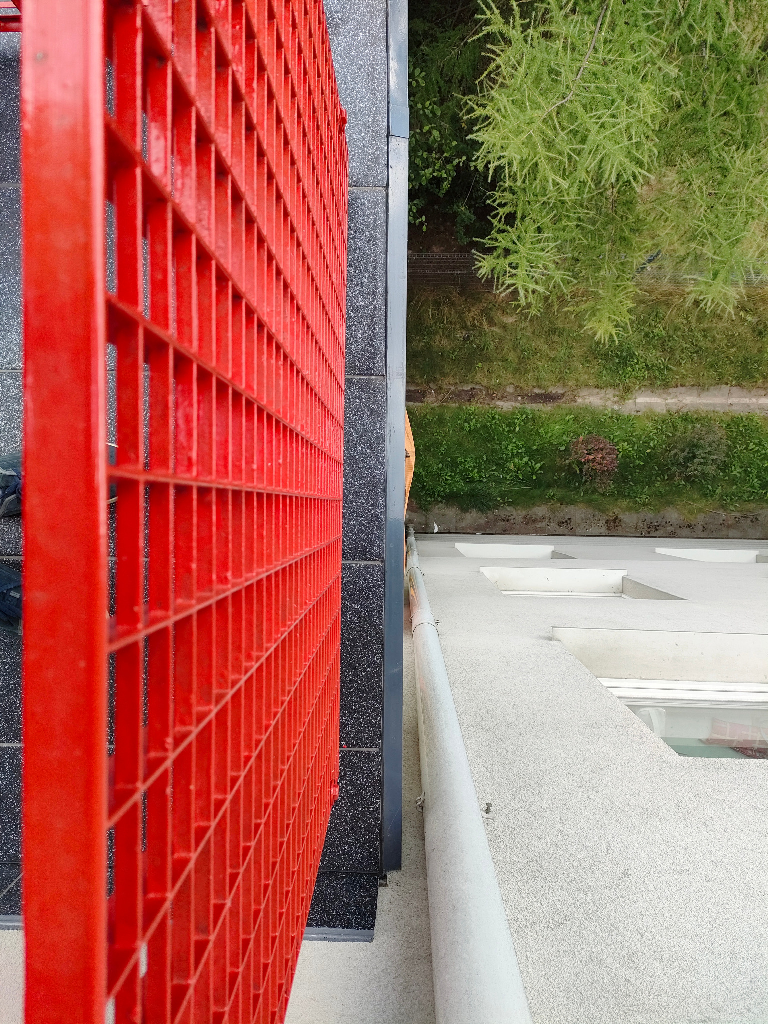Czerwona, metalowa balustrada balkonu z widokiem na zieleń i elewację budynku. Nietypowy kadr z góry, podkreślający geometrię i kontrast kolorów.