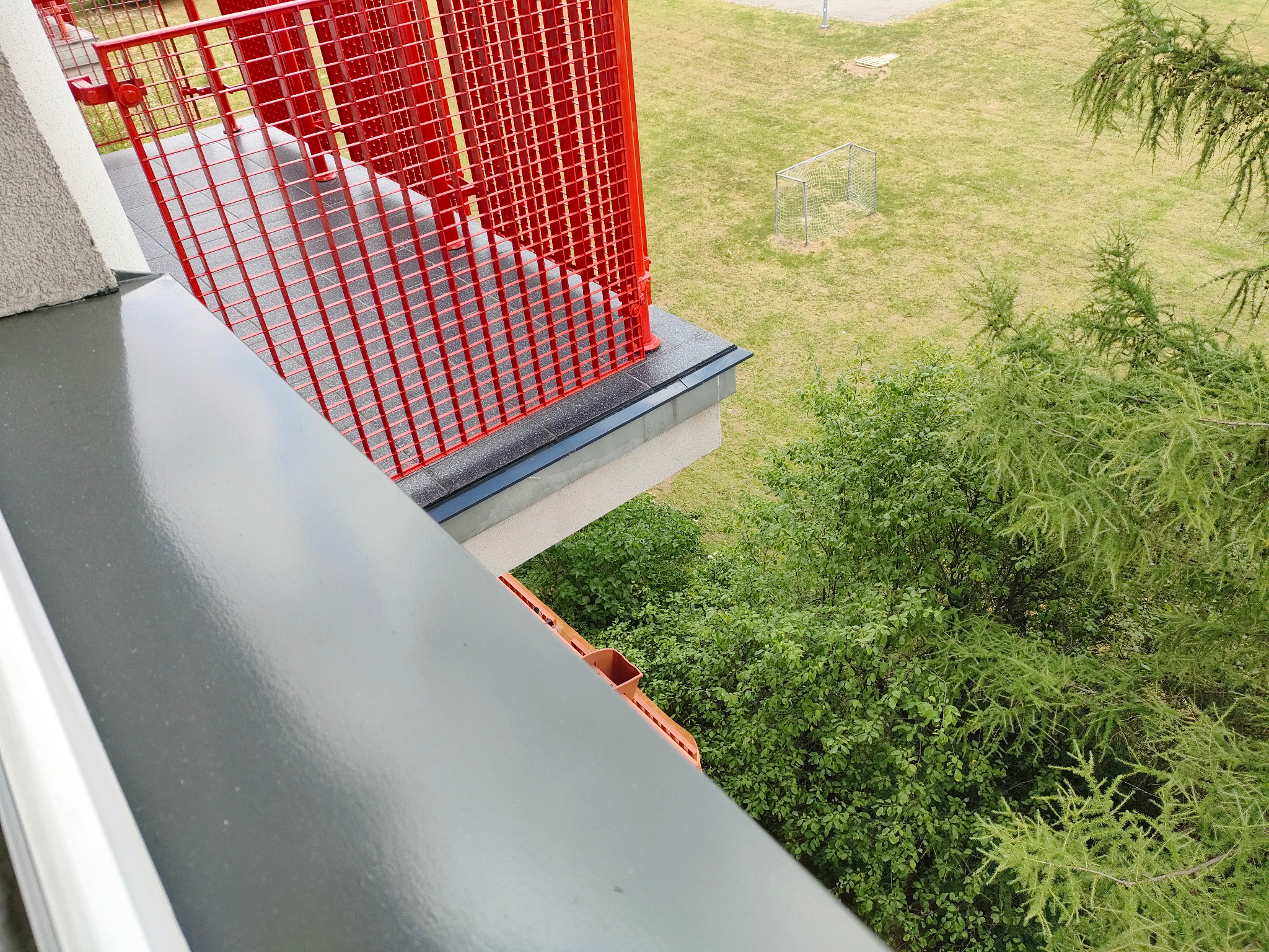 Balkon z czerwoną, metalową balustradą widziany z góry, z widokiem na zieleń i boisko. Szara, gładka powierzchnia balkonu w pierwszym planie.