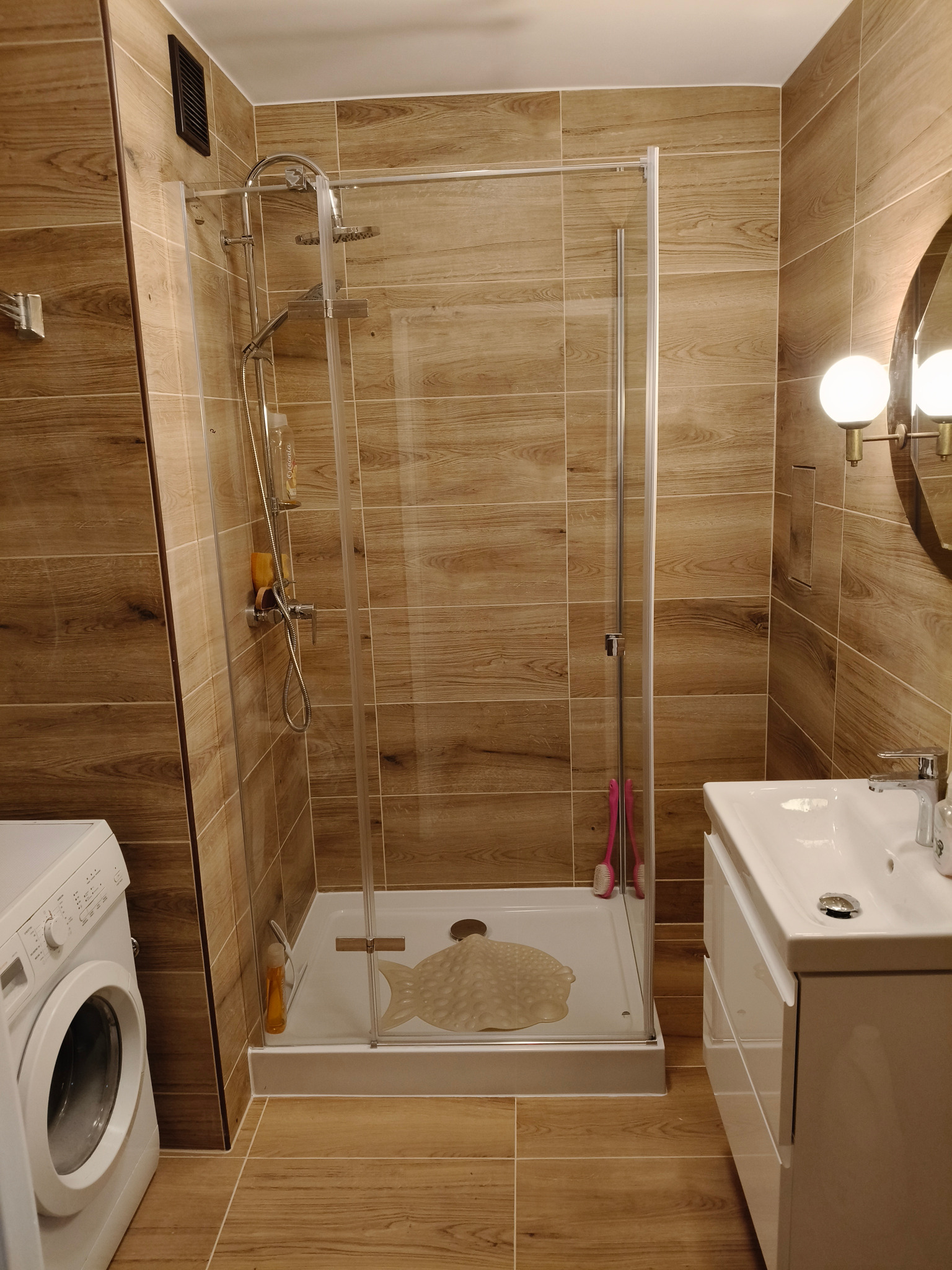 Nowoczesna łazienka z kabiną prysznicową typu walk-in, umywalką z szafką i pralką. Płytki imitujące drewno na ścianach i podłodze. Oświetlenie punktowe.
