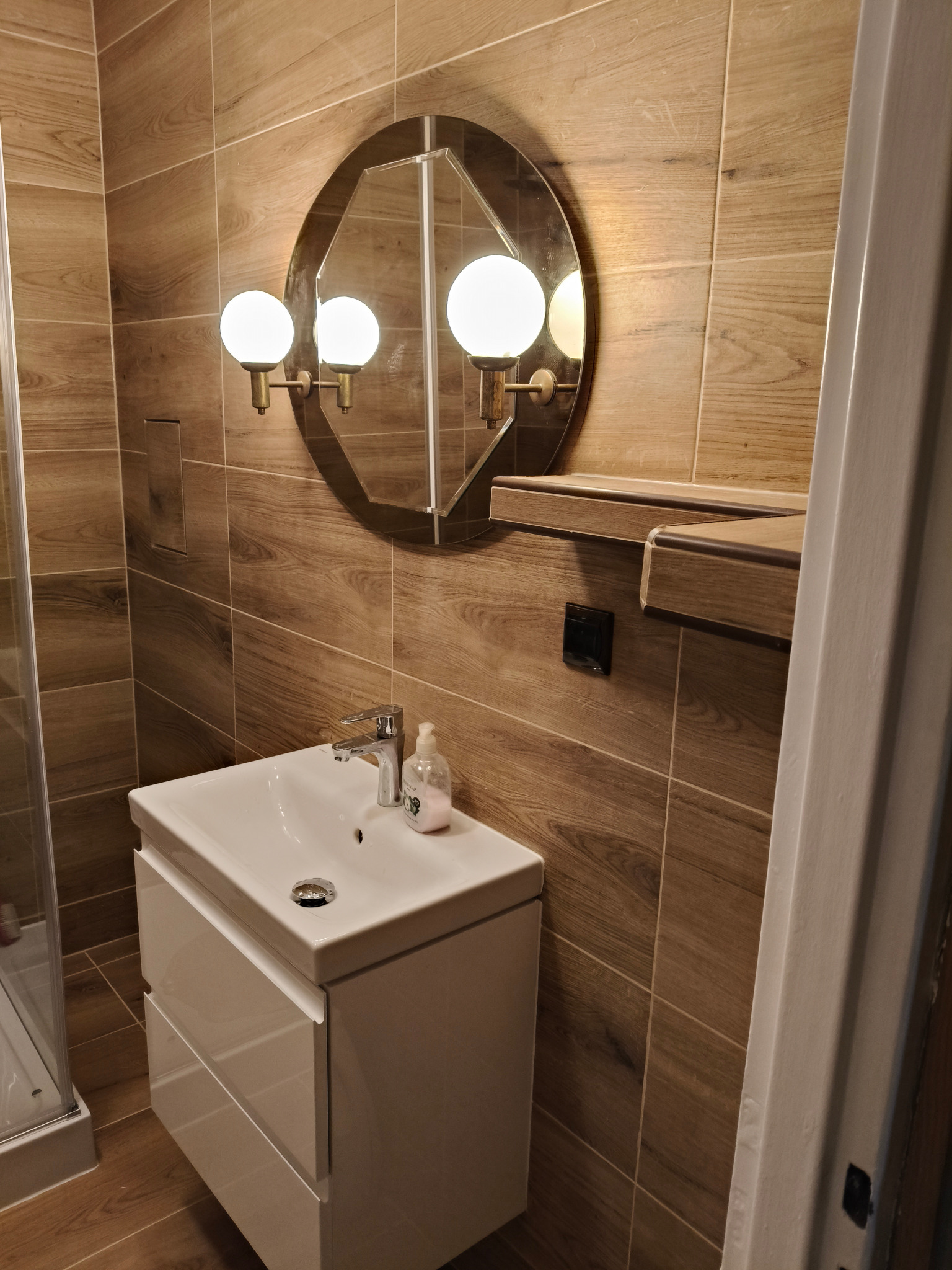 Nowoczesna łazienka z drewnopodobnymi płytkami, owalnym lustrem z trzema kulistymi lampami, białą szafką pod umywalką i kabiną prysznicową w tle.