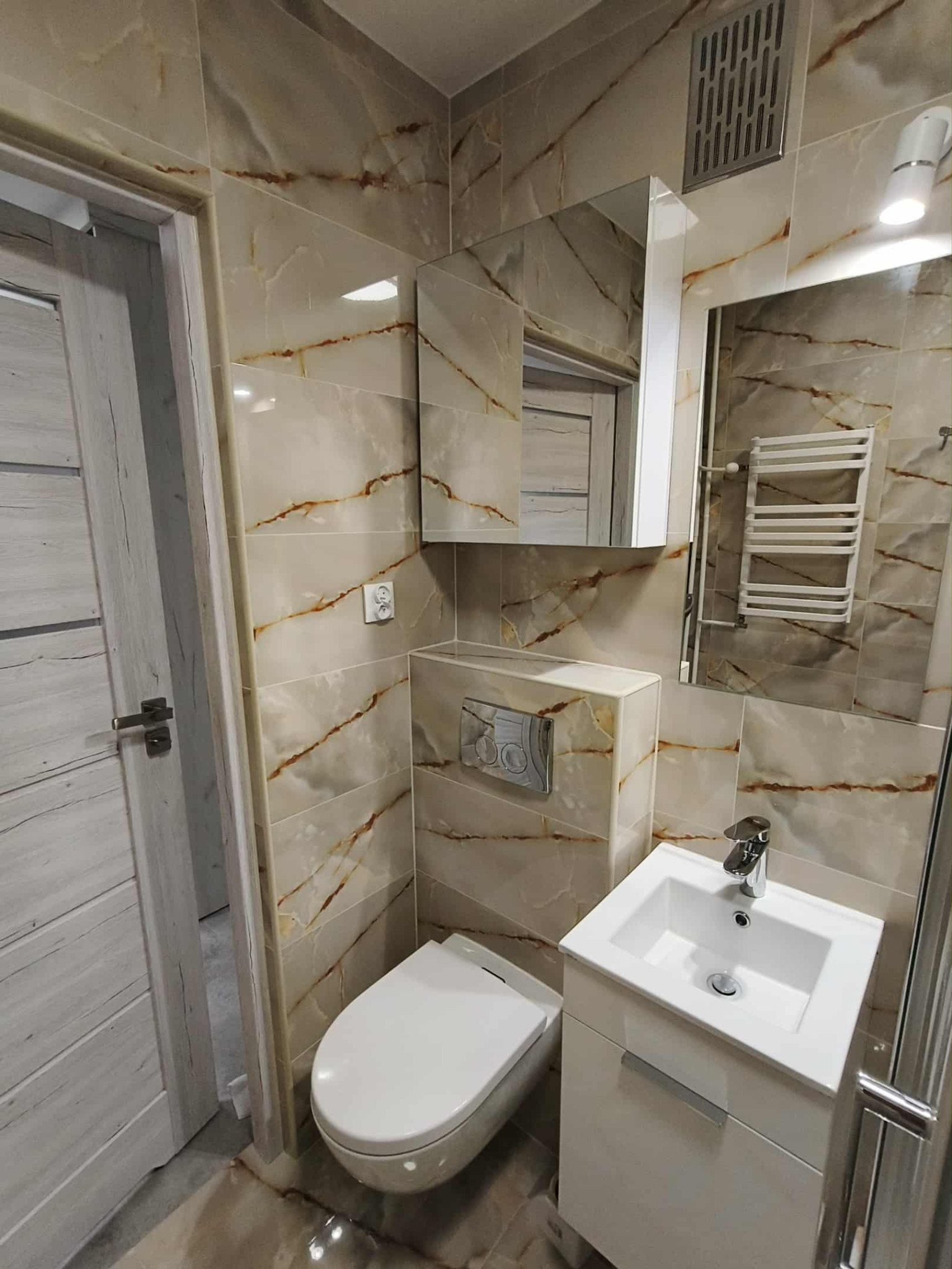 Wykończona łazienka w Krakowie z beżowymi płytkami imitującymi marmur, białą umywalką z szafką i toaletą podwieszaną. Widoczny grzejnik drabinkowy i lustro.