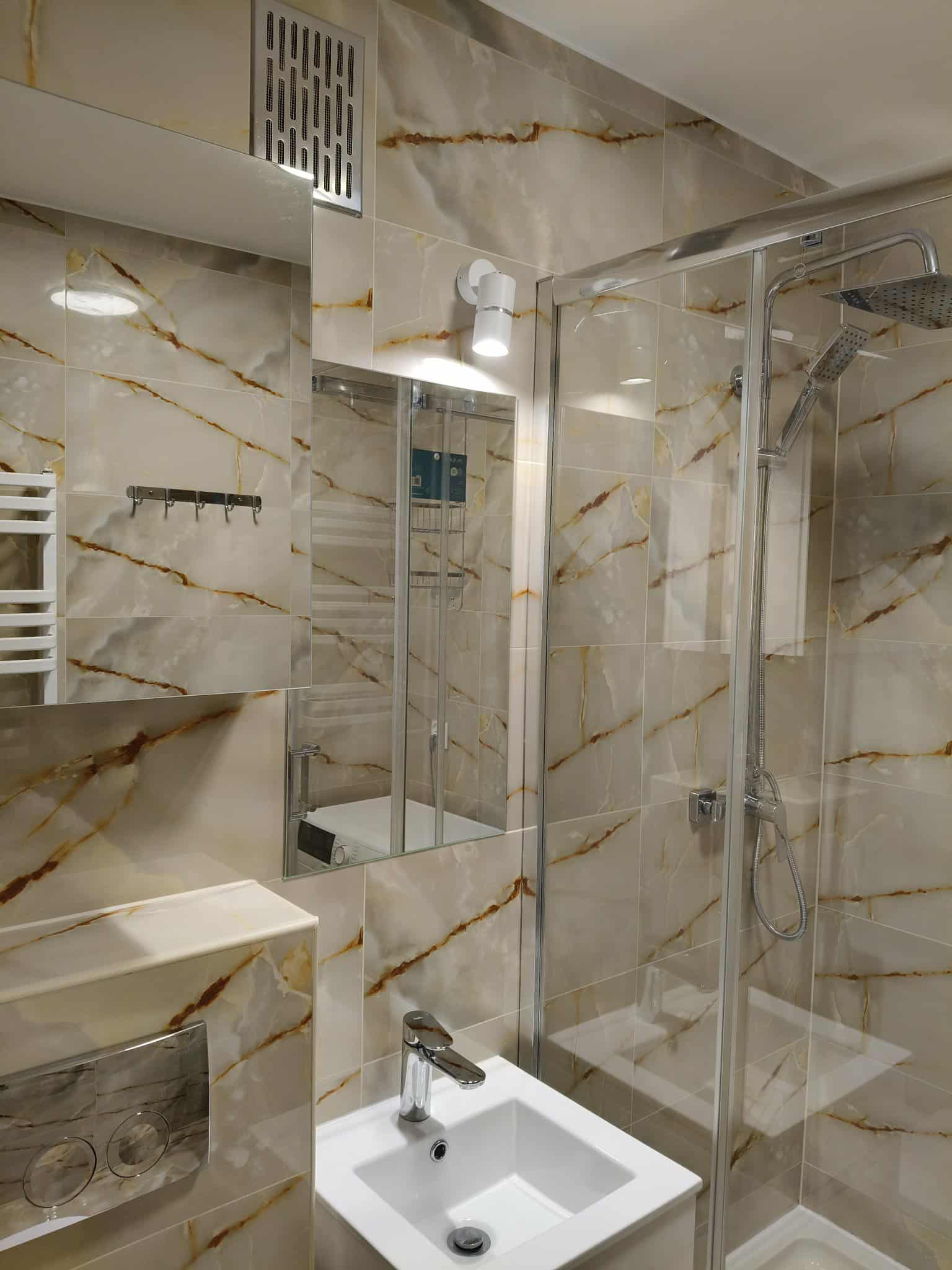 Wykończona łazienka z kabiną prysznicową, umywalką i grzejnikiem. Płytki imitujące marmur z brązowymi żyłami. Nowoczesna armatura i oświetlenie.