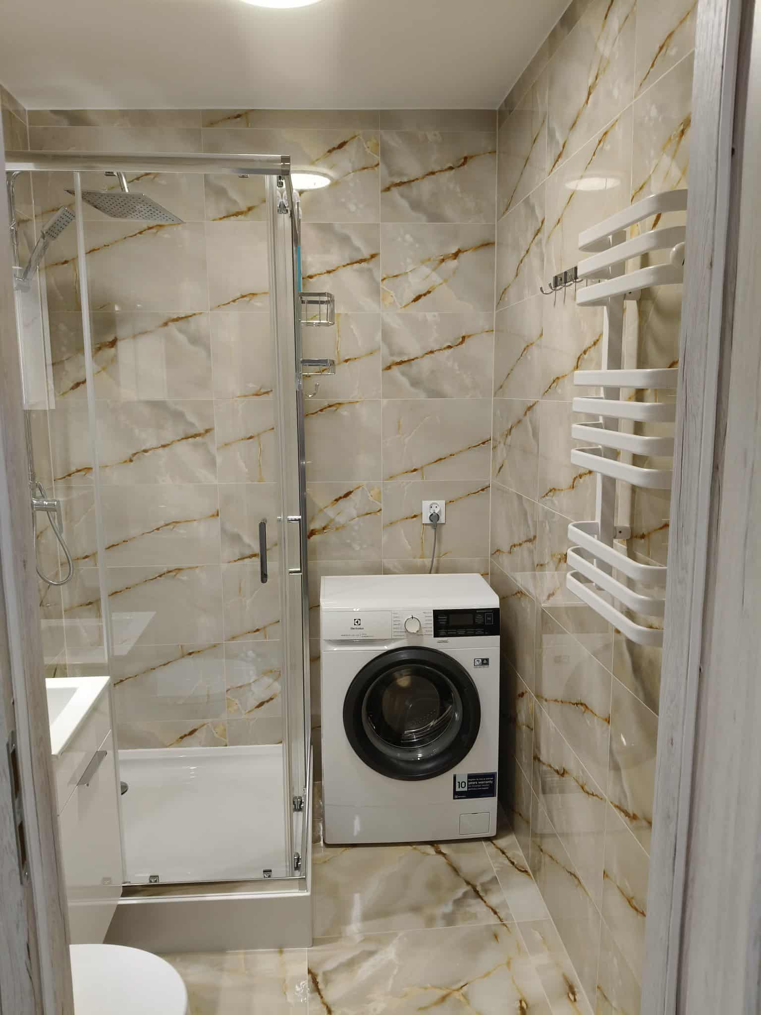Wykończona łazienka z kabiną prysznicową, pralką i grzejnikiem drabinkowym na tle marmurowych płytek. Widok na wnętrze łazienki z perspektywy wejścia.