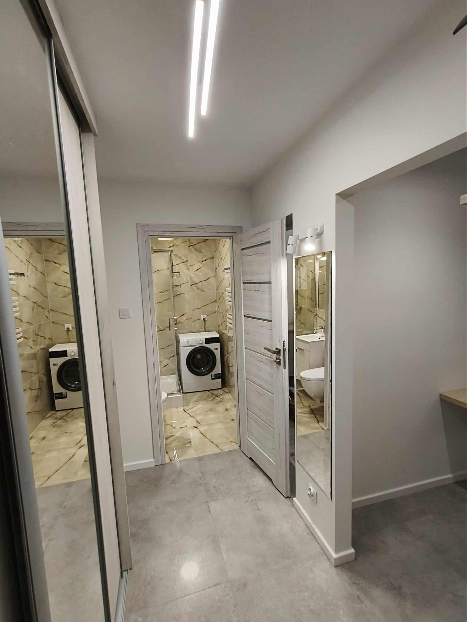 Wykończona łazienka z pralką i marmurowymi płytkami, widoczna z korytarza przez otwarte drzwi. W korytarzu szafa z lustrem i wejście do drugiej łazienki.