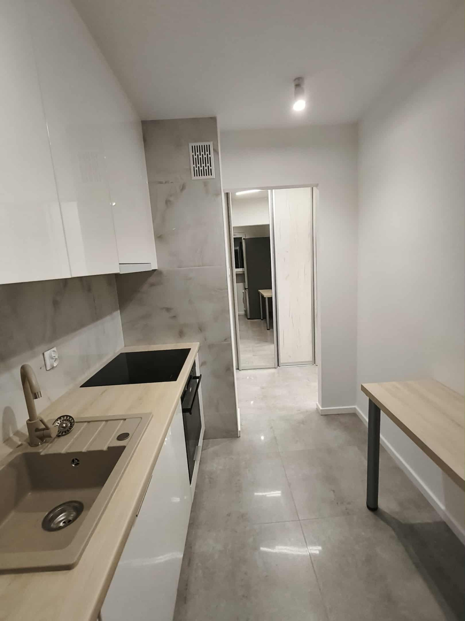 Nowoczesna kuchnia z beżowym zlewem i białymi szafkami, wykończona płytkami imitującymi marmur. Widok na korytarz z przesuwnymi drzwiami.