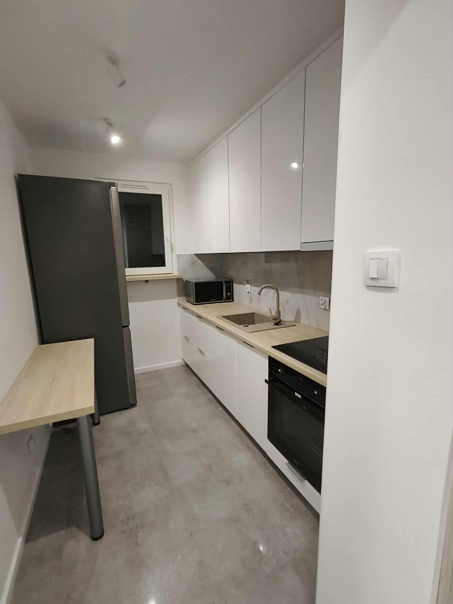 Nowoczesna, wąska kuchnia z białymi, błyszczącymi szafkami, szarą lodówką i blatem w odcieniach drewna. Widoczny zlew, płyta indukcyjna i piekarnik. Minimalistyczny design.