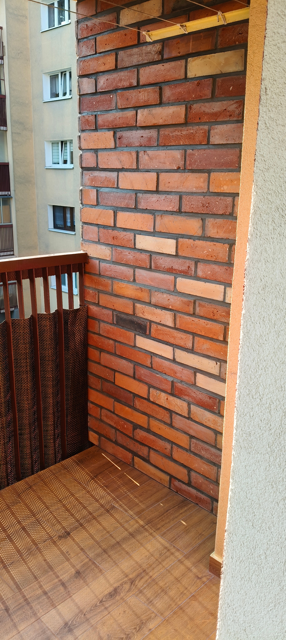 Ceglana ściana na balkonie z drewnianą podłogą i osłoną. Widok na budynek mieszkalny w tle. Słoneczne światło podkreśla teksturę cegieł i podłogi.
