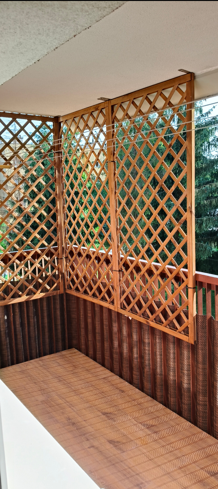 Zadaszony balkon z drewnianą kratą i brązową okładziną ścian. Widok na zieleń przez kratę. Ukończona realizacja wykończenia balkonu.