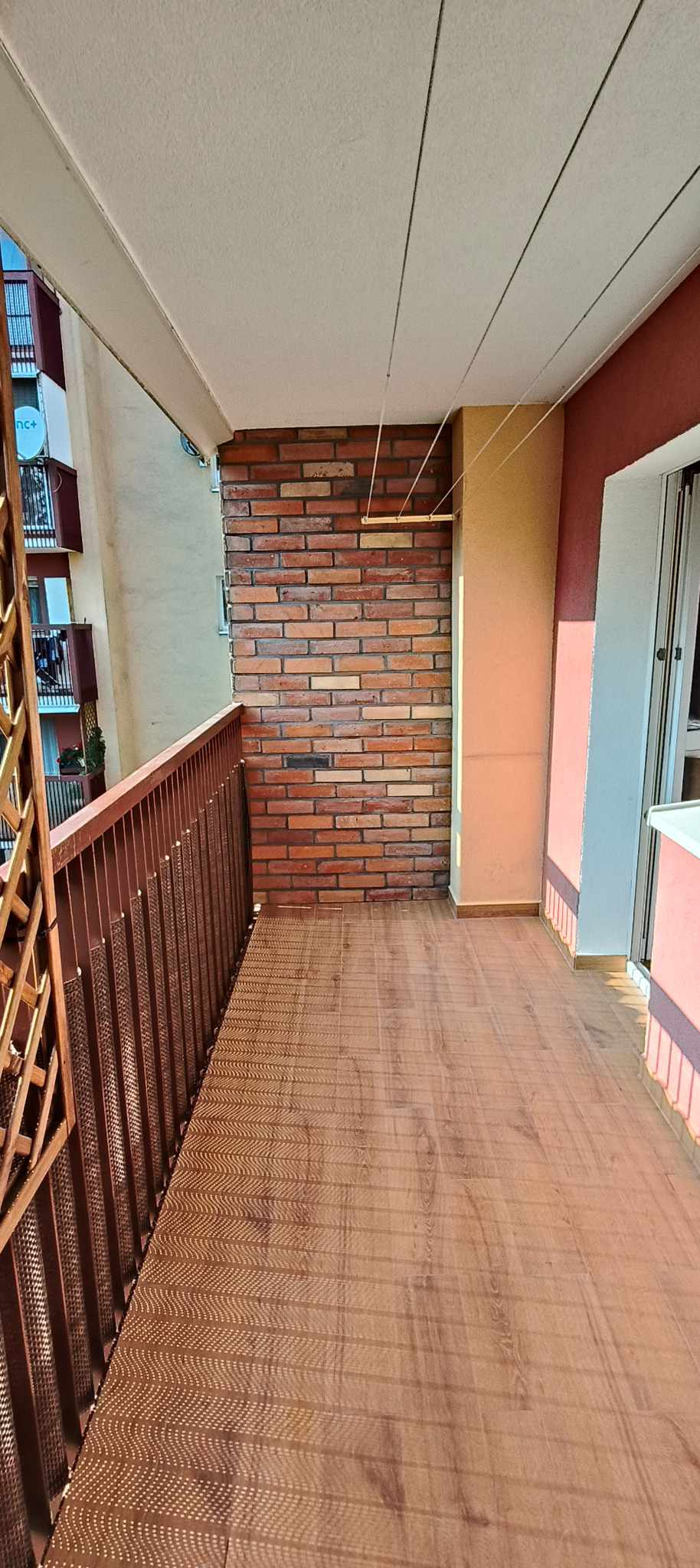 Wykończony balkon z drewnopodobną podłogą i ścianą z cegły. Widoczne brązowe barierki i fragment budynku w tle. Uwagę przykuwa nietypowy wzór na podłodze.