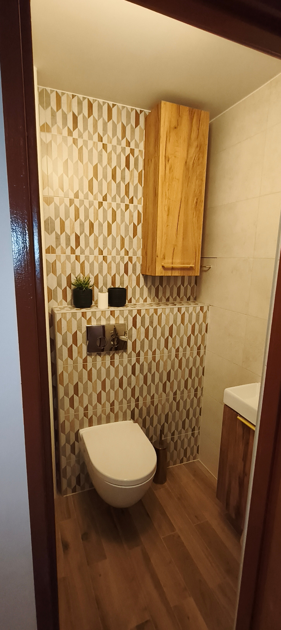 Nowoczesna toaleta z geometrycznymi płytkami w odcieniach beżu i brązu, drewnianą szafką wiszącą i podłogą imitującą drewno. Uwagę przykuwają detale, takie jak rośliny w doniczkach.