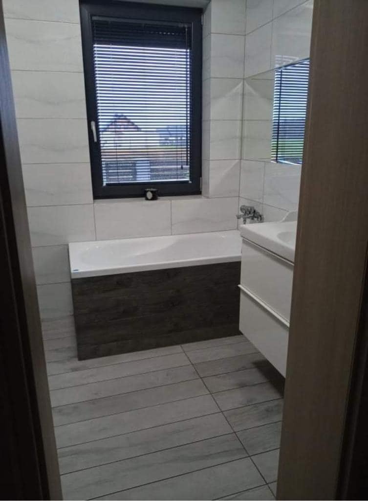 Nowoczesna łazienka z biało-szarymi płytkami, wanną z ciemnobrązową obudową imitującą drewno, białą umywalką z szafką i czarnym oknem z żaluzjami.