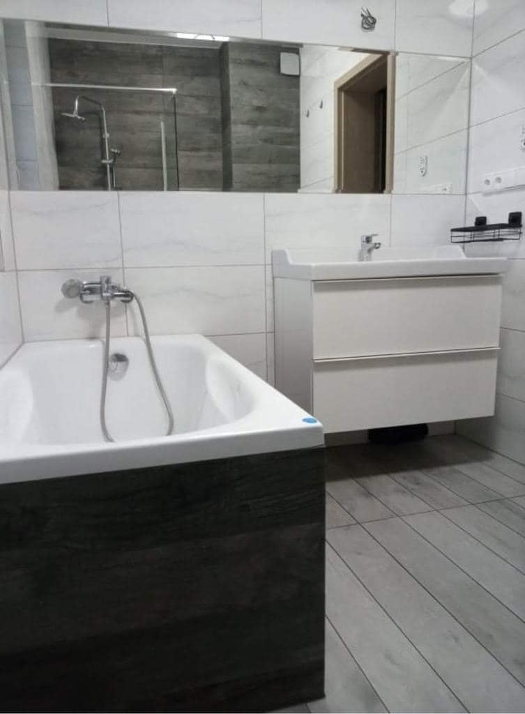 Nowoczesna łazienka z wanną obudowaną ciemnym drewnem, białą umywalką z szafką i dużym lustrem odbijającym kabinę prysznicową.